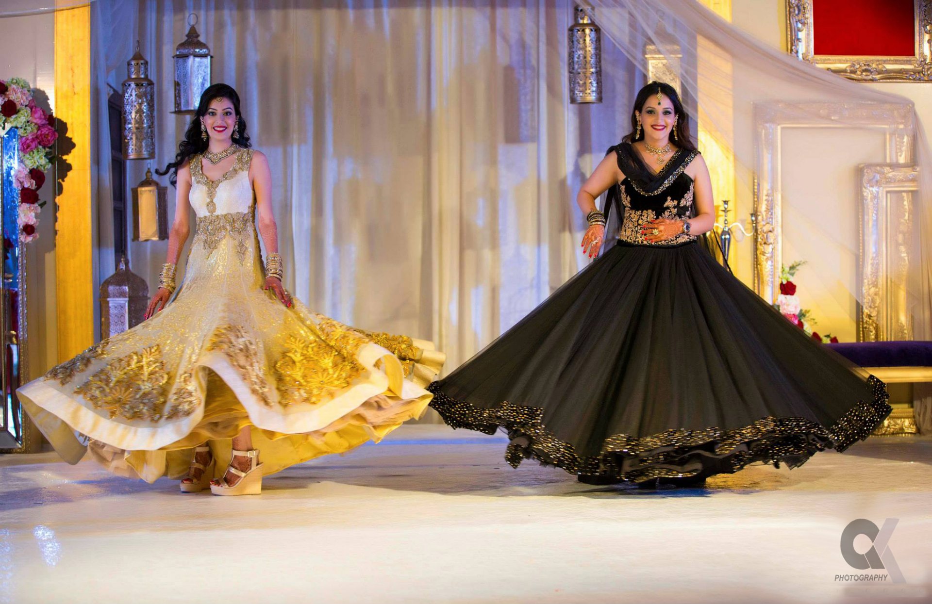 Two women twirling in flowing gold and black gowns on a decorated stage.