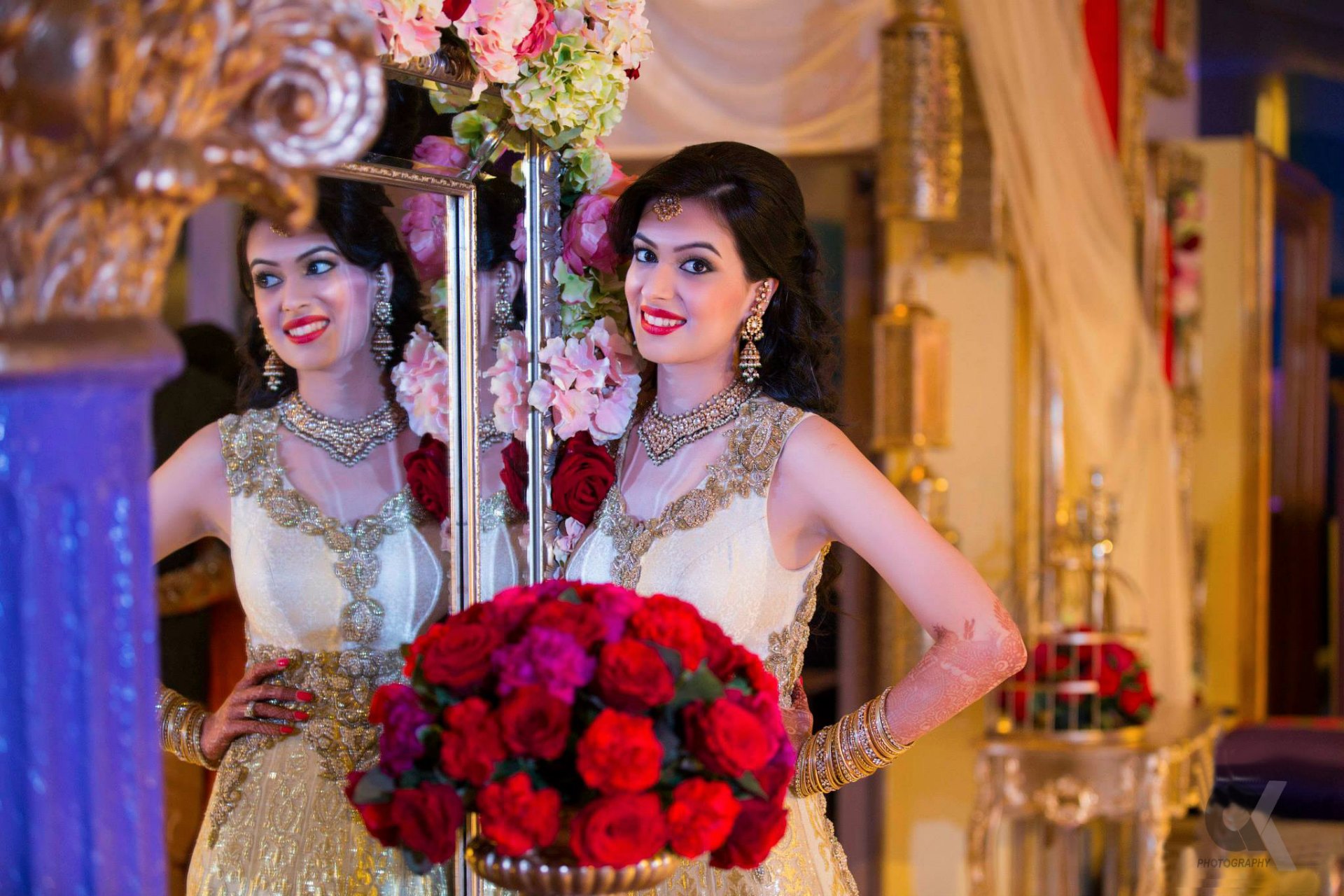 A bride in ornate attire posing beside a mirror with red roses.