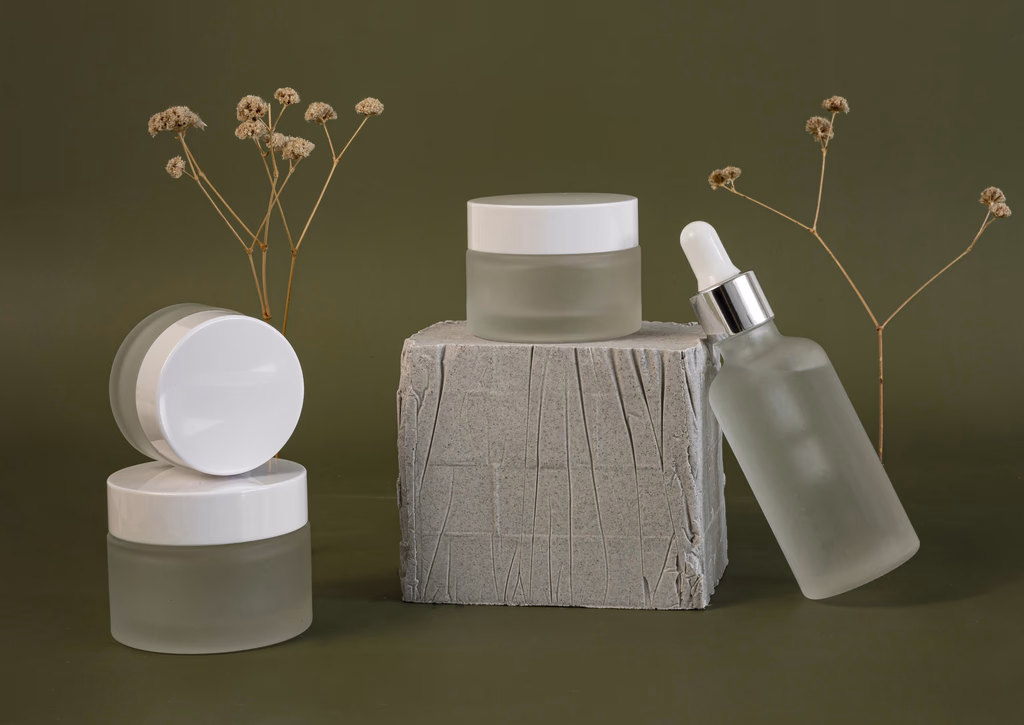 Minimal skincare bottles and jars arranged with dried botanicals against a deep green background.